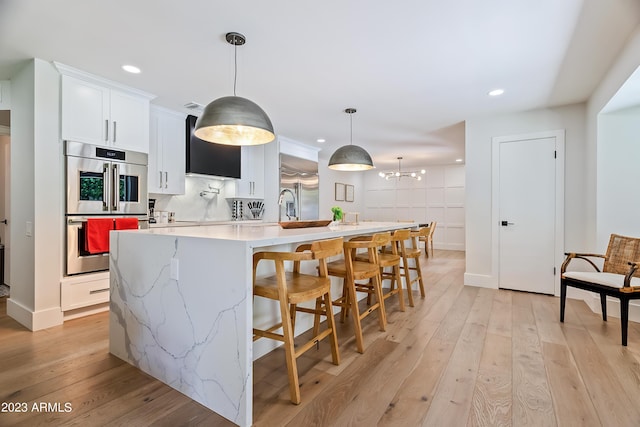 kitchen with white cabinets, a center island with sink, light wood-type flooring, decorative light fixtures, and stainless steel double oven