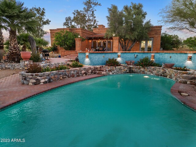 pool at dusk featuring pool water feature and a patio