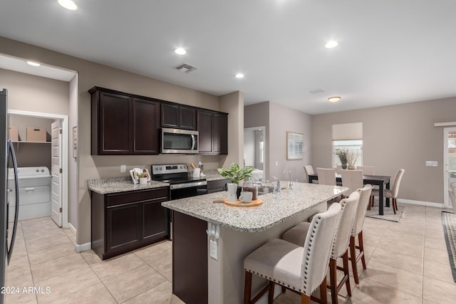 kitchen with light stone counters, dark brown cabinetry, stainless steel appliances, light tile patterned floors, and an island with sink