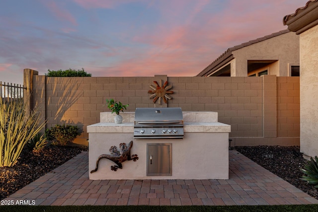 patio terrace at dusk featuring an outdoor kitchen, a fenced backyard, and grilling area