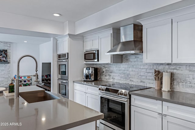 kitchen featuring decorative backsplash, appliances with stainless steel finishes, white cabinets, a sink, and wall chimney range hood