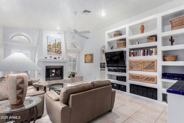 living room with light tile patterned floors, visible vents, a fireplace with raised hearth, a ceiling fan, and built in shelves