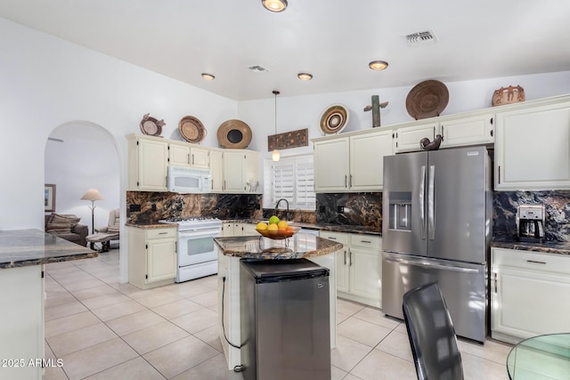 kitchen featuring arched walkways, light tile patterned flooring, white appliances, a center island, and dark stone counters