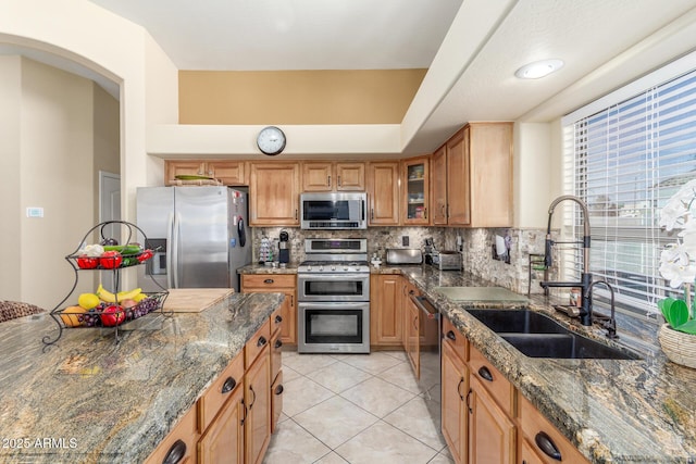 kitchen featuring a sink, stainless steel appliances, tasteful backsplash, dark stone counters, and glass insert cabinets