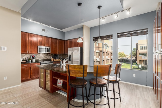 kitchen featuring tasteful backsplash, hanging light fixtures, dark stone countertops, a center island with sink, and appliances with stainless steel finishes