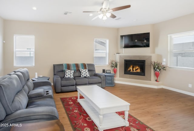 living room with visible vents, light wood-style flooring, a tiled fireplace, ceiling fan, and baseboards