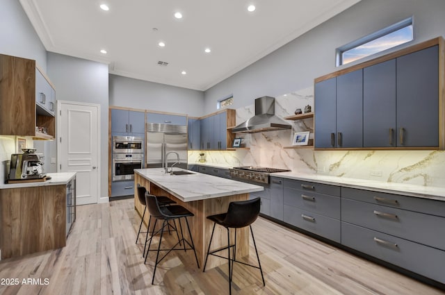 kitchen featuring open shelves, an island with sink, a breakfast bar area, wall chimney range hood, and appliances with stainless steel finishes