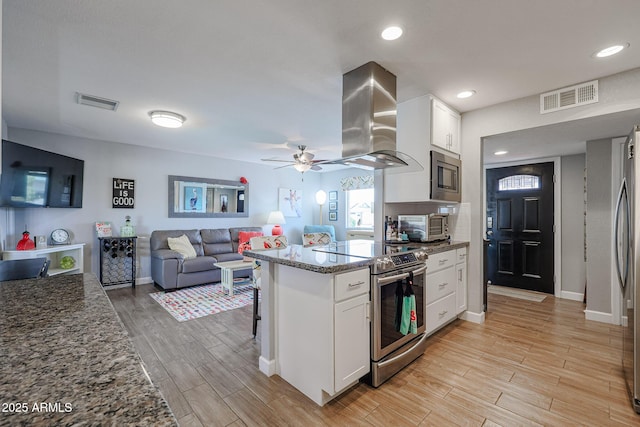 kitchen featuring appliances with stainless steel finishes, island range hood, white cabinetry, dark stone counters, and light wood-type flooring