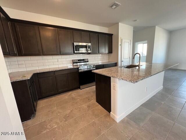 kitchen with an island with sink, sink, backsplash, light stone counters, and stainless steel appliances