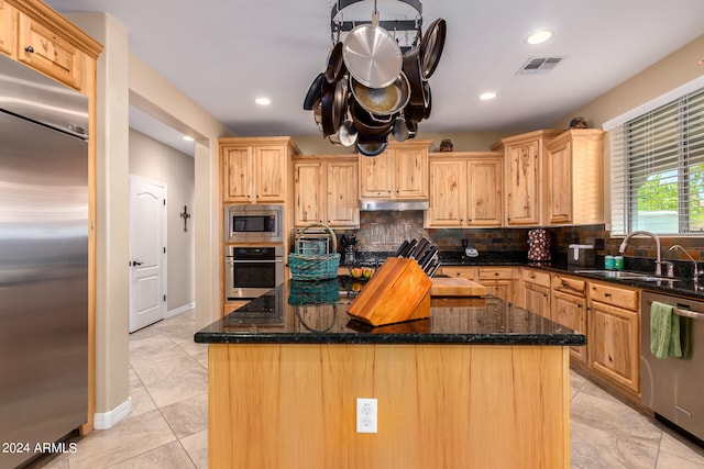 kitchen with dark stone countertops, a kitchen island, light brown cabinets, built in appliances, and sink