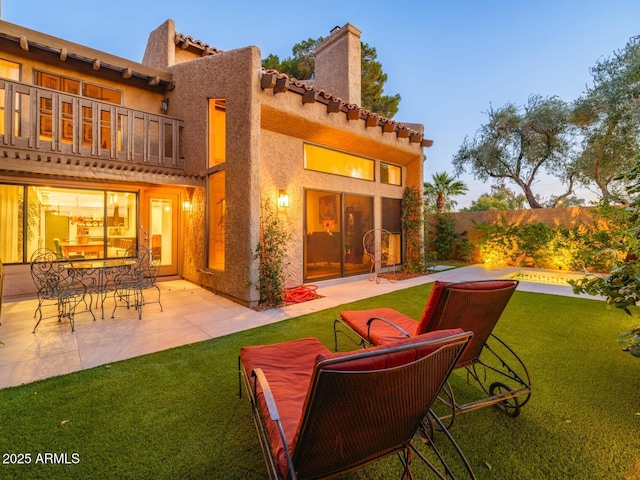 back house at dusk with a balcony, a patio area, and a lawn