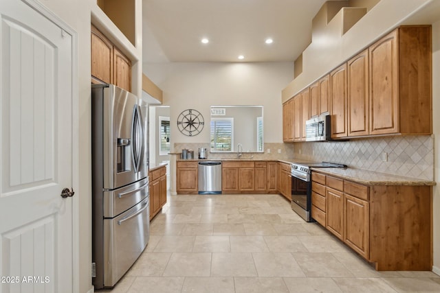 kitchen featuring light stone countertops, decorative backsplash, brown cabinets, appliances with stainless steel finishes, and a sink