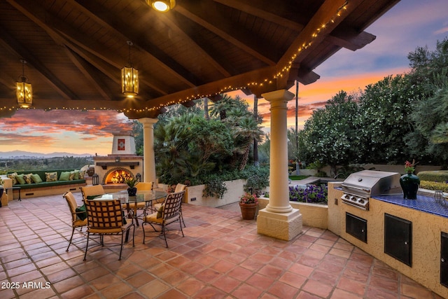 patio terrace at dusk with grilling area, a lit fireplace, a gazebo, outdoor dining area, and area for grilling