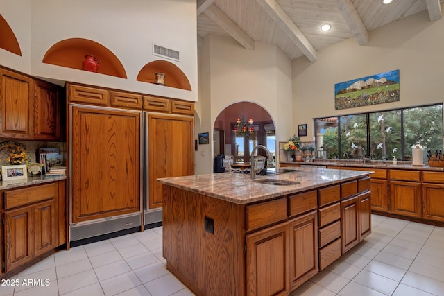 kitchen with brown cabinetry, visible vents, light stone countertops, a sink, and beamed ceiling