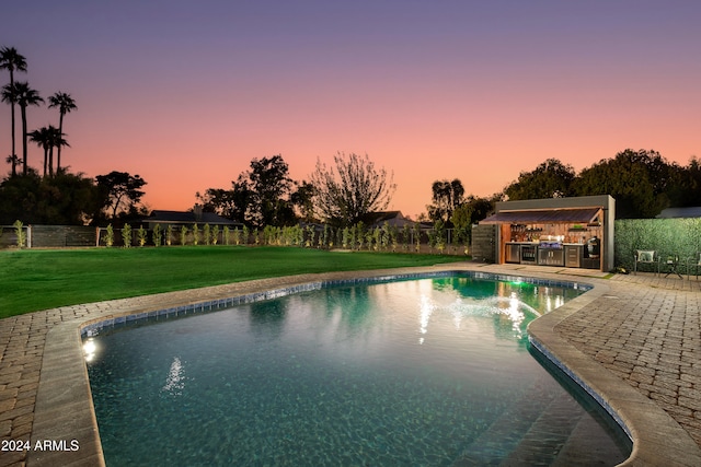 pool at dusk with exterior kitchen, a patio area, and a yard