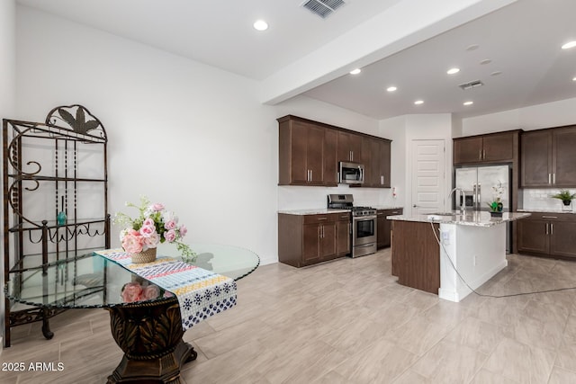 kitchen featuring appliances with stainless steel finishes, beamed ceiling, a kitchen island with sink, light stone counters, and dark brown cabinetry