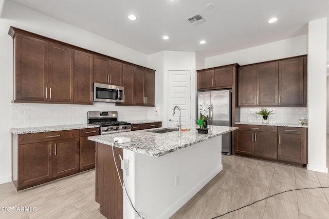 kitchen featuring dark brown cabinetry, sink, a center island with sink, appliances with stainless steel finishes, and light stone countertops