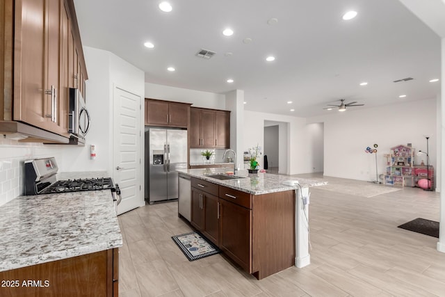 kitchen with sink, ceiling fan, appliances with stainless steel finishes, light stone counters, and an island with sink