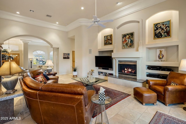 tiled living room with built in shelves, ceiling fan, a tile fireplace, decorative columns, and crown molding