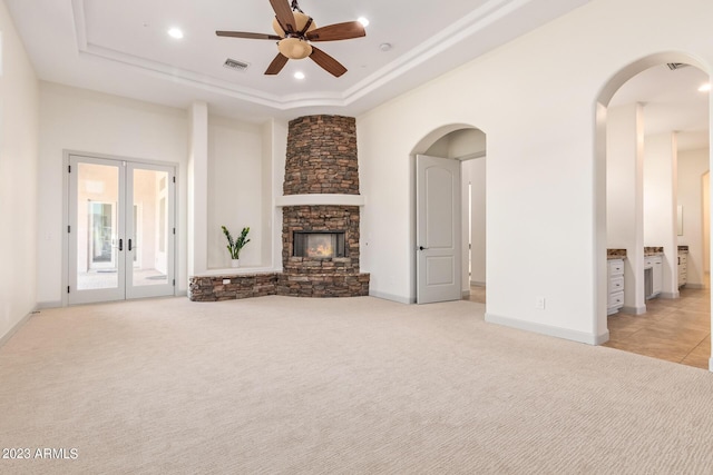 unfurnished living room featuring light carpet, french doors, a raised ceiling, a stone fireplace, and ceiling fan