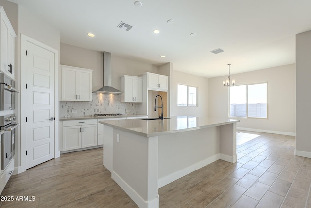 kitchen featuring white cabinetry, sink, wall chimney exhaust hood, decorative backsplash, and a center island with sink