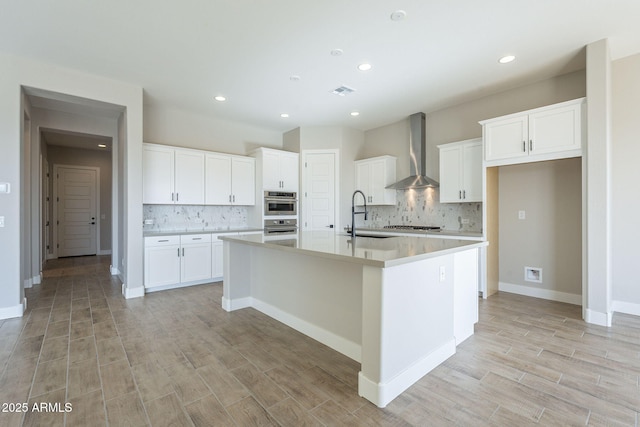 kitchen with sink, an island with sink, white cabinetry, and wall chimney range hood