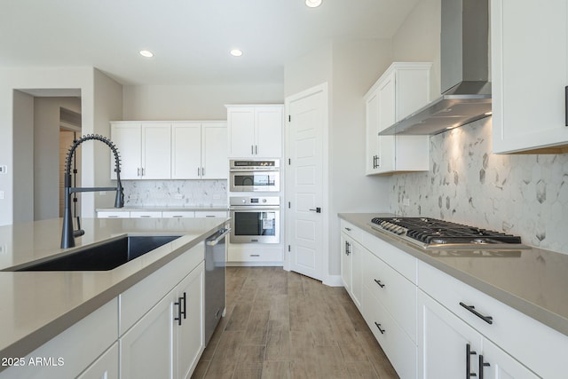 kitchen with appliances with stainless steel finishes, wall chimney exhaust hood, sink, light hardwood / wood-style flooring, and white cabinetry