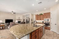 kitchen with ceiling fan, sink, light stone counters, an island with sink, and oven