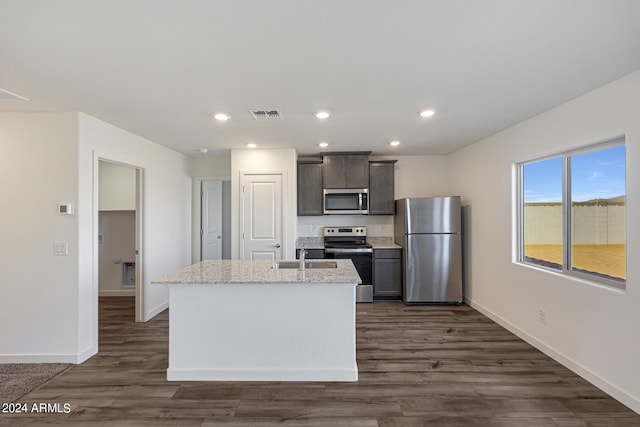 kitchen featuring stainless steel appliances, an island with sink, light stone countertops, and dark wood-type flooring