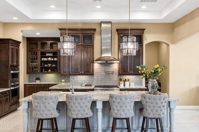 bar featuring dark brown cabinetry, wall chimney range hood, decorative light fixtures, and tasteful backsplash