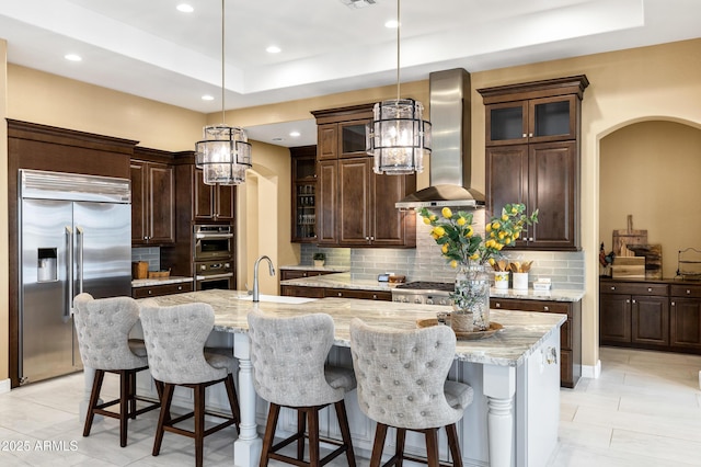 kitchen featuring decorative light fixtures, sink, stainless steel appliances, a center island with sink, and wall chimney range hood