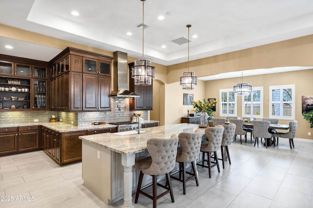 kitchen featuring wall chimney exhaust hood, dark brown cabinets, a center island with sink, a tray ceiling, and light stone countertops