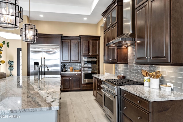 kitchen featuring dark brown cabinetry, wall chimney exhaust hood, high end appliances, pendant lighting, and light stone countertops