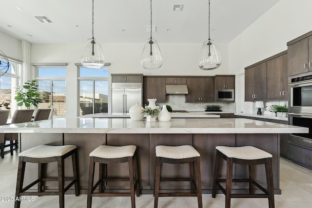 20475 E Indiana Ave, Queen Creek, AZ 85142 - kitchen with under cabinet range hood, built in appliances, dark brown cabinets, a large island, and tasteful backsplash