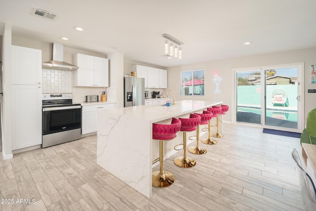 kitchen with pendant lighting, stainless steel appliances, wall chimney range hood, a kitchen island with sink, and white cabinets