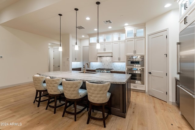 kitchen with light stone counters, stainless steel appliances, decorative light fixtures, light hardwood / wood-style flooring, and an island with sink