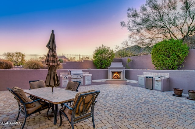 patio terrace at dusk with a grill, exterior fireplace, and exterior kitchen