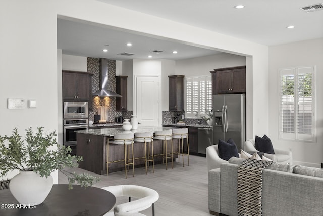 kitchen featuring stainless steel appliances, visible vents, open floor plan, dark brown cabinets, and wall chimney exhaust hood