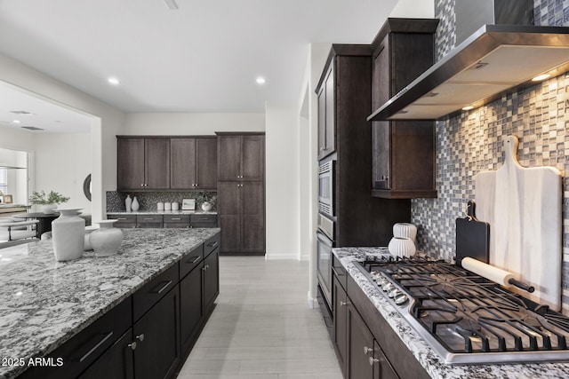 kitchen with light stone countertops, dark brown cabinets, appliances with stainless steel finishes, wall chimney range hood, and decorative backsplash