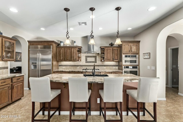 kitchen with wall chimney exhaust hood, stainless steel appliances, backsplash, an island with sink, and decorative light fixtures