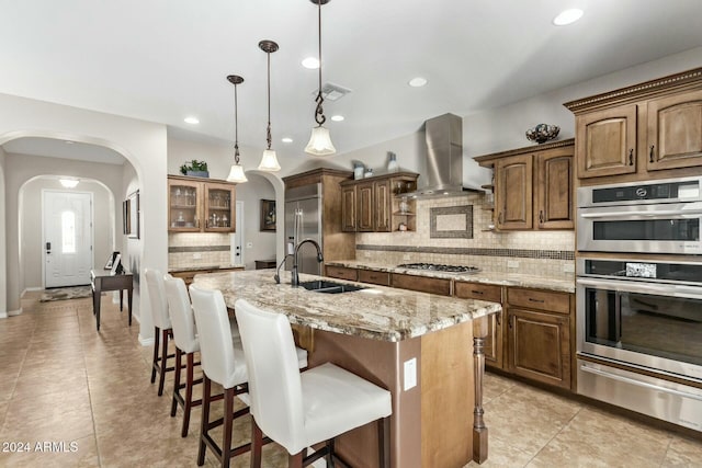 kitchen with ventilation hood, sink, light stone countertops, an island with sink, and appliances with stainless steel finishes