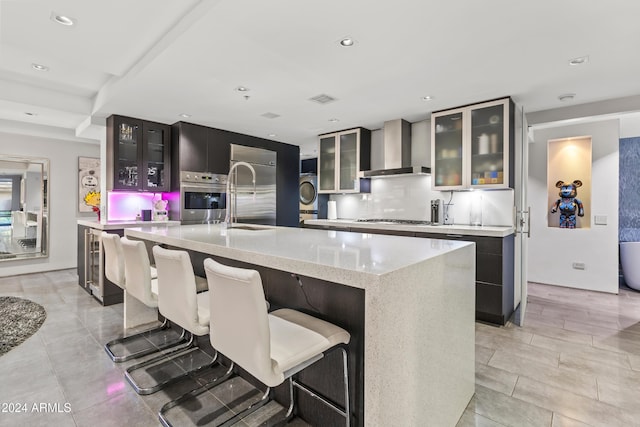 kitchen with wall chimney range hood, stainless steel appliances, a kitchen island with sink, and tasteful backsplash