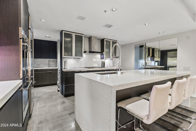 kitchen featuring sink, wall chimney exhaust hood, an island with sink, a kitchen bar, and appliances with stainless steel finishes