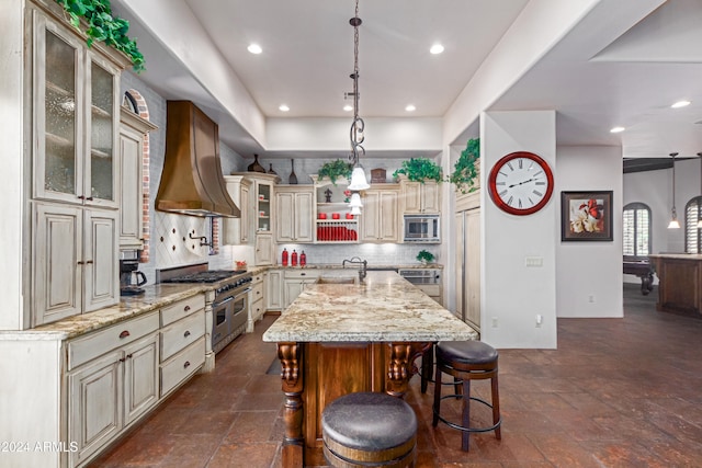 kitchen featuring backsplash, custom exhaust hood, stainless steel appliances, a kitchen island with sink, and a breakfast bar area