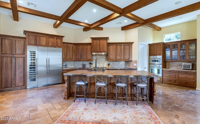 kitchen featuring appliances with stainless steel finishes, a kitchen island with sink, a kitchen breakfast bar, coffered ceiling, and beamed ceiling