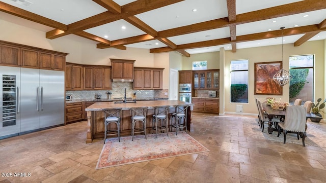 kitchen with beamed ceiling, an island with sink, a breakfast bar area, a high ceiling, and stainless steel appliances