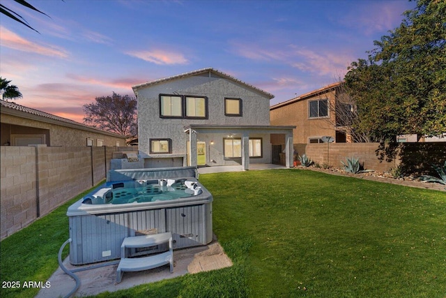 back house at dusk with a hot tub, a patio area, and a lawn