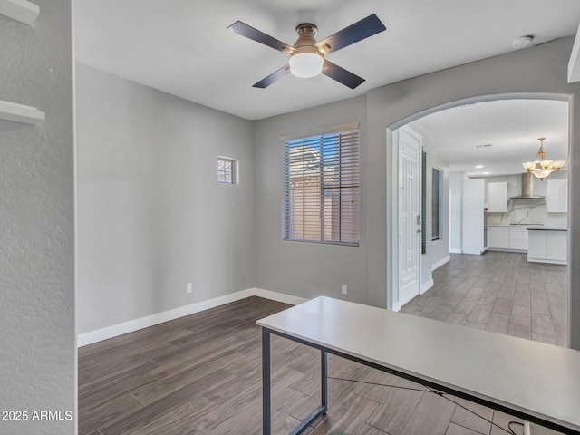 unfurnished office featuring arched walkways, ceiling fan with notable chandelier, wood finished floors, and baseboards