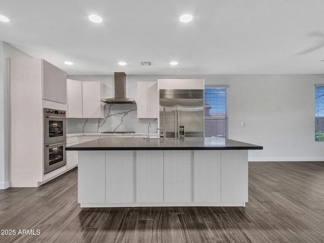 kitchen featuring dark wood-style flooring, tasteful backsplash, dark countertops, appliances with stainless steel finishes, and wall chimney range hood