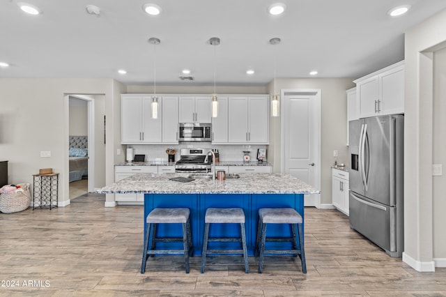 kitchen with light wood-type flooring, a kitchen island with sink, stainless steel appliances, white cabinets, and hanging light fixtures